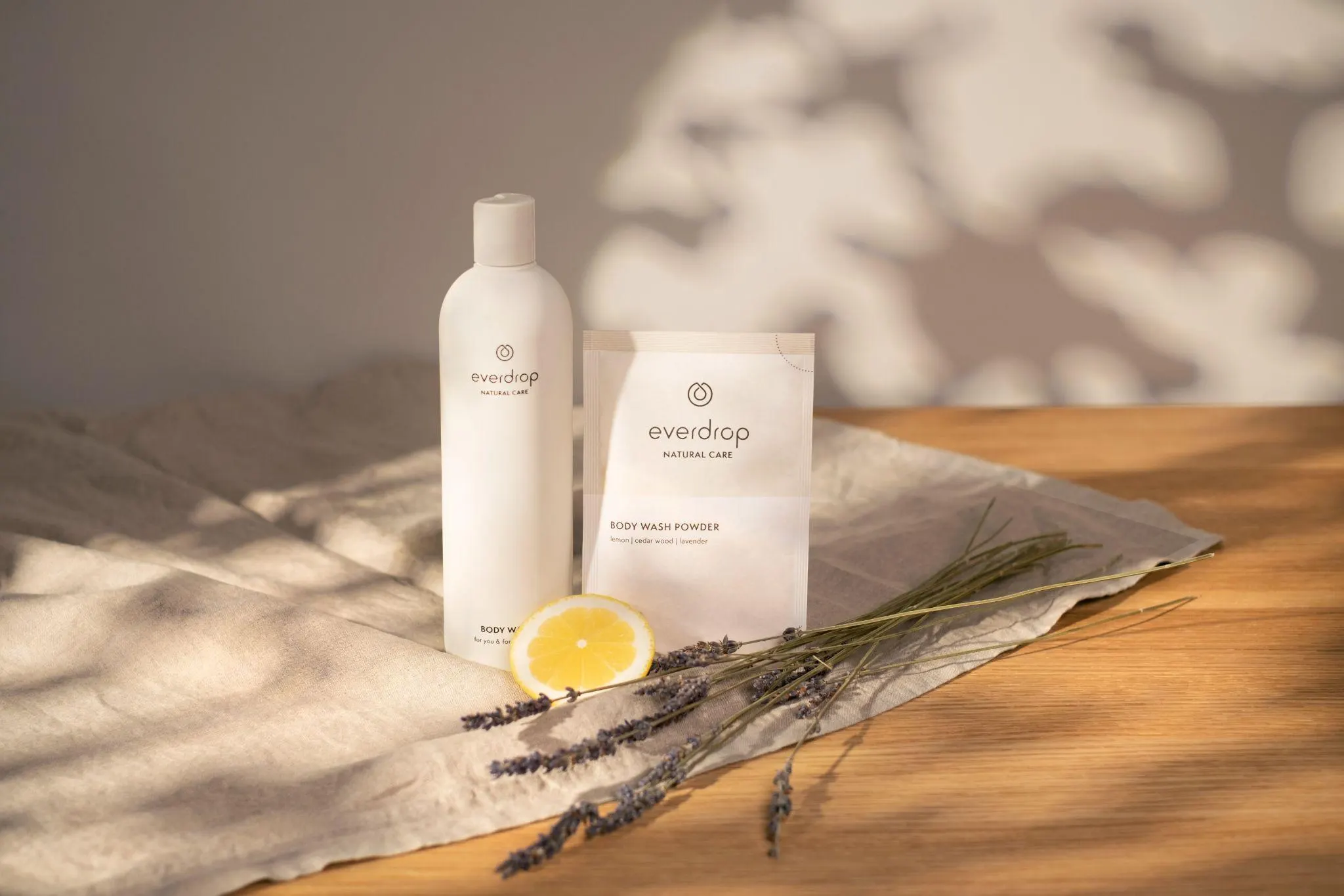 Produktaufnahme im Tageslicht: everdrop-Body-Wash-Flasche und Body-Wash-Powder-Sachet auf Leinentuch, daneben eine Zitronenscheibe und getrockneter Lavendel