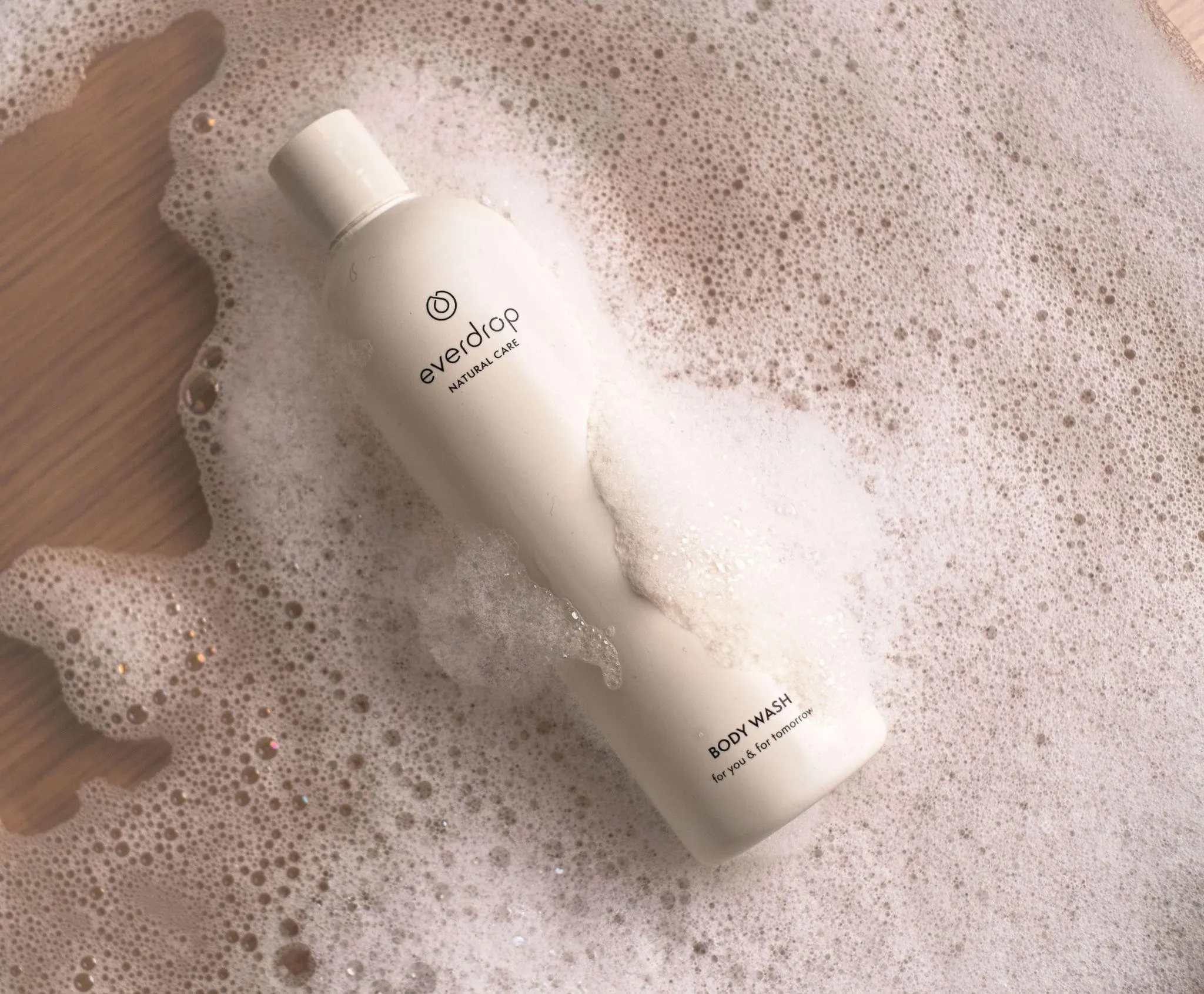 Aufsicht auf eine everdrop-Body-Wash-Flasche, die auf einer Holzfläche in Seifenschaum liegt